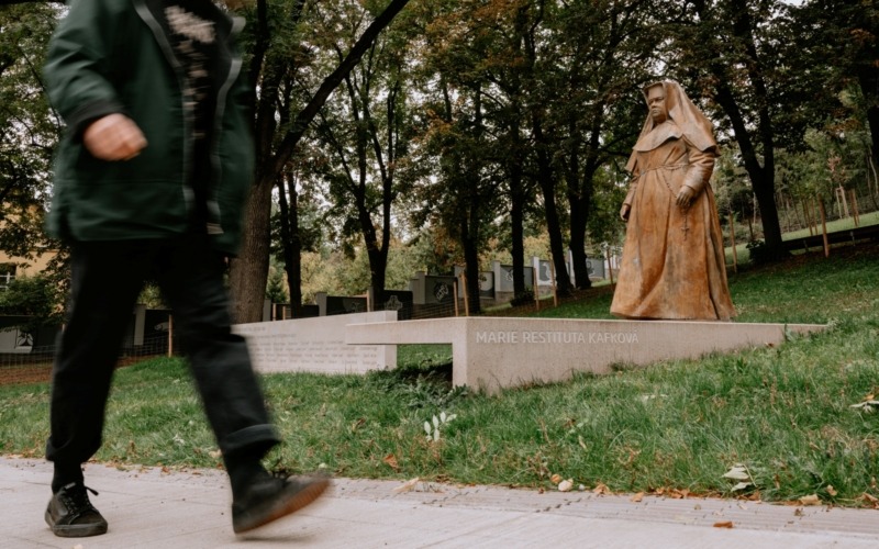 Socha Marie Restituty na betonovém podstavci s pamětní stěnou v pozadí.