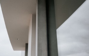 Sloup z pohledového betonu s jemnou strukturou u prosklené fasády polyfunkčního domu.